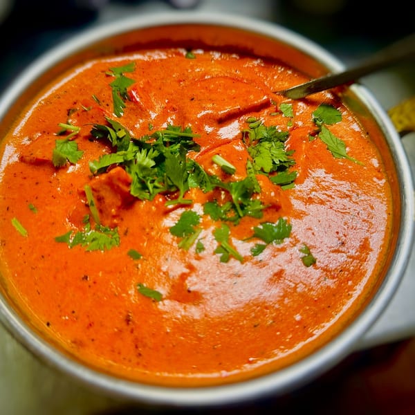 Close-up of a vibrant orange Indian curry, garnished with fresh green cilantro, in a metal bowl.