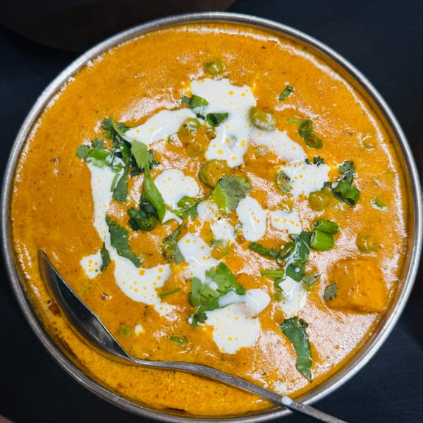 Close-up of a vibrant orange Indian curry with peas, paneer, cilantro, and cream in a metal bowl.